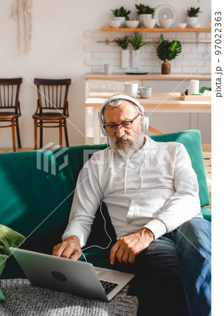 Handsome old man dressed in hoodie and eyeglasses is using laptop and smiling while listen music sitting on couch at home Handsome old man dressed in hoodie and eyeglasses is using laptop and smiling while listen music sitting on couch at home 97022363