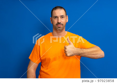 Bearded handsome hispanic man wearing orange casual t-shirt standing over isolated blue background doing happy thumbs up gesture with hand. Approval expression looking at camera showing success 97023858