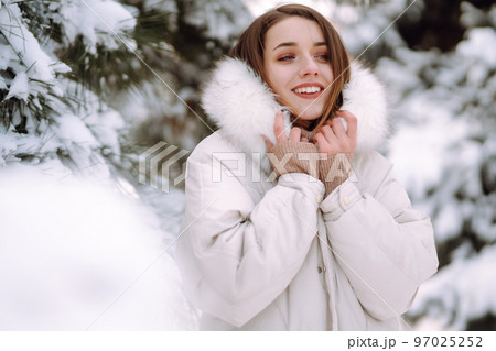 Beautiful woman in winter clothes posing in a snowy park. Young lady walking in a sunny winter day. 97025252