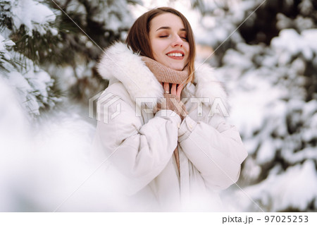 Beautiful woman in winter clothes posing in a snowy park. Young lady walking in a sunny winter day. 97025253