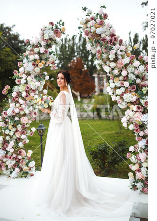 Beautiful fiancee in white wedding dress with bouquet stand near the wedding arch of flowers. Beautiful fiancee in white wedding dress with bouquet stand near the wedding arch of flowers. 97027021