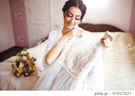 Morning of beautiful bride near the wedding dress. Bride in a white robe holds and admires her wedding dress. 97027027