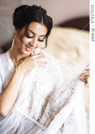Morning of beautiful bride near the wedding dress. Bride in a white robe holds and admires her wedding dress. 97027035