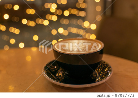 A cup of latte coffee with heart-shaped foam on a table in a cafe, in the background, the lights in a blur are reflected on the table. 97027773