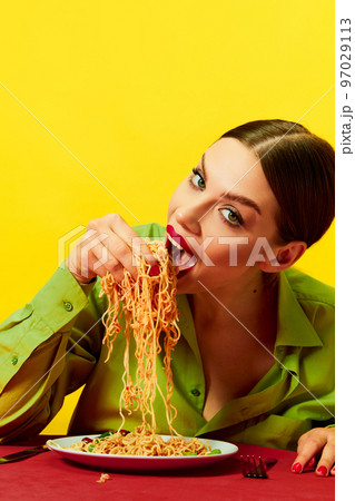 Emotional young girl eating spaghetti, noodles with hands on red tablecloth over yellow background. Feeling hungry. Food pop art photography. Emotional young girl eating spaghetti, noodles with hands on red tablecloth over yellow background. Feeling hungry. Food pop art photography. 97029113