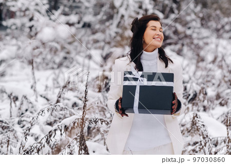A stylish woman with a white suit with a New Year's gift in her hands in a winter forest. A girl in nature in a snowy forest with a gift box 97030860