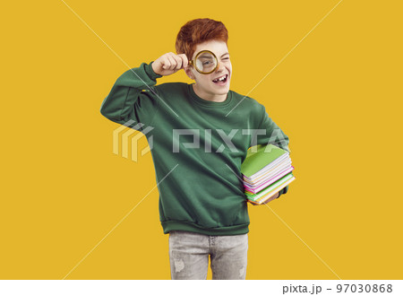 School boy standing on yellow background, holding books and looking through magnifying glass 97030868