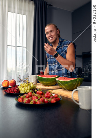 Handsome young sporty smiling man in the kitchen is preparing vegan healthy fruits salad and smoothie in a good mood 97032160
