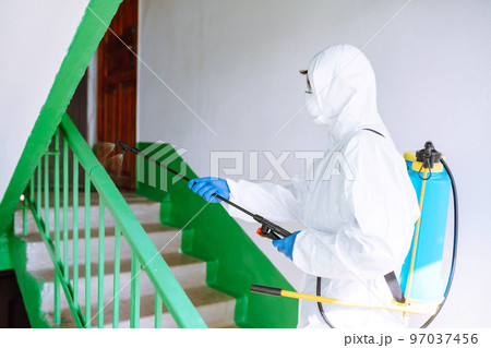 Man in Hazmat suit, protective gloves and goggles use sprayer equipment disinfect the entrance. Man in Hazmat suit, protective gloves and goggles use sprayer equipment disinfect the entrance. 97037456