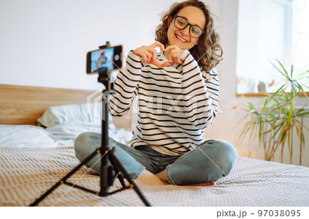 Young woman blogger talking with followers, live streaming, looking to phone screen sitting on bed. 97038095