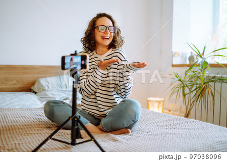 Young woman blogger talking with followers, live streaming, looking to phone screen sitting on bed. 97038096