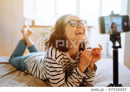 Young woman blogger talking with followers, live streaming, looking to phone screen sitting on bed. 97038097