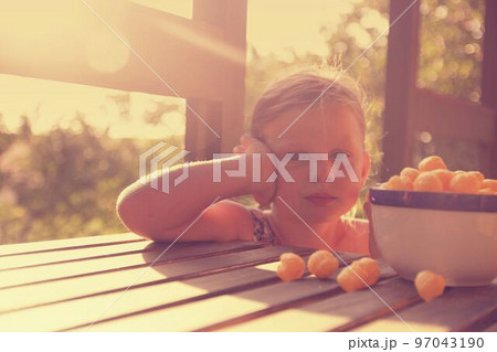 Little girl is sitting on porch in summer. Cheese balls in bowl. Cheese flavoured snacks on a table. Dreamy and romantic image. Summer and happy childhood concept Little girl is sitting on porch in summer. Cheese balls in bowl. Cheese flavoured snacks on a table. Dreamy and romantic image. Summer and happy childhood concept 97043190