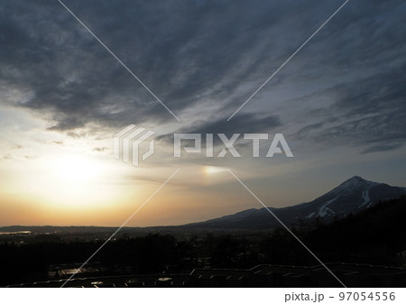 早春、冠雪の磐梯山の麓に光る夕日、オレンジ色の一部に虹色の雲が出現し雄大な景色が印象的 97054556
