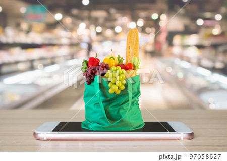 Fresh food and vegetables in green shopping bag on mobile smartphone on wood table with supermarket aisle blurred background grocery online concept 97058627