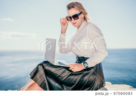 Digital nomad, Business woman working on laptop by the sea. Pretty lady typing on computer by the sea at sunset, makes a business transaction online from a distance. Freelance, remote work on vacation 97060083