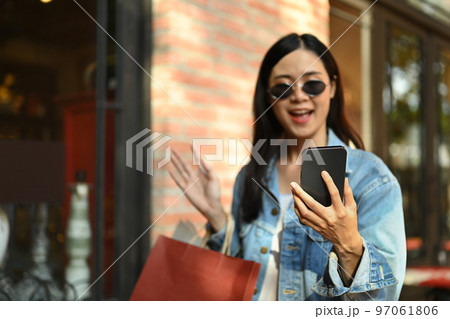 Cheerful asian woman taking selfie or making video call on mobile phone while standing at the shopping district of a city 97061806