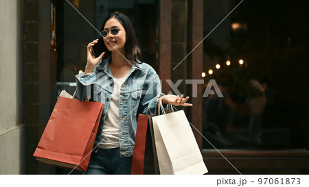 Confident stylish woman with shopping bags talking on mobile phone while standing near showcase in city street 97061873