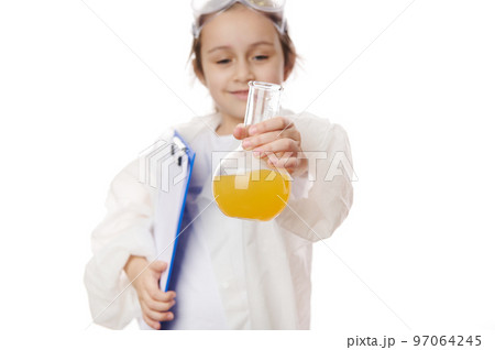 Selective focus on laboratory flask with yellow liquid chemical, in hands of smart elementary school student in lab coat, holding clipboard, showing chemical reaction taking place in flask to camera Selective focus on laboratory flask with yellow liquid chemical, in hands of smart elementary school student in lab coat, holding clipboard, showing chemical reaction taking place in flask to camera 97064245