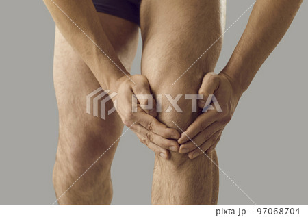 Close up of hands of man holding and massaging sore knee standing on gray background. Close up of hands of man holding and massaging sore knee standing on gray background. 97068704