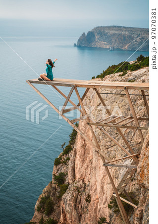 A girl sits on a wooden springboard for jumping with a rope. In a dark green dress with her hands in the air. A girl sits on a wooden springboard for jumping with a rope. In a dark green dress with her hands in the air. 97070381