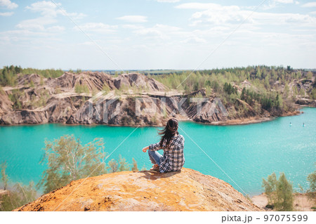Young woman sitting on the top of mounting and feeling free and looking at the blue lake Young woman sitting on the top of mounting and feeling free and looking at the blue lake 97075599