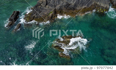 Black moss-covered rocks among turquoise waves of the Celtic Sea. White sea foam on the waves. Waters of the Atlantic Ocean. Aerial photo. Black moss-covered rocks among turquoise waves of the Celtic Sea. White sea foam on the waves. Waters of the Atlantic Ocean. Aerial photo. 97077842