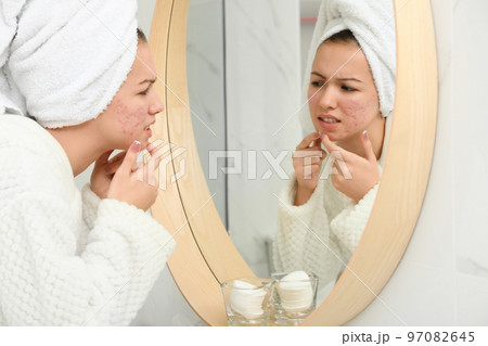 Teen girl with acne problem squeezing pimple near mirror in bathroom 97082645