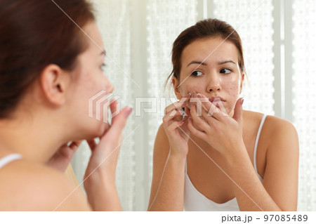 Teen girl with acne problem squeezing pimple near mirror in bathroom Teen girl with acne problem squeezing pimple near mirror in bathroom 97085489
