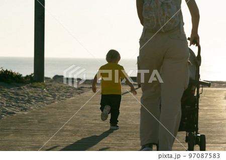 A woman drives a baby stroller and her little son runs forward to the ocean A woman drives a baby stroller and her little son runs forward to the ocean 97087583
