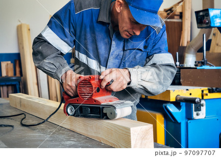 Carpenter in work clothes processes wood with grinder. Polishing of wood surface. Middle-aged man 97097707