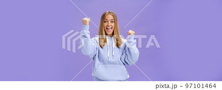 Cheerful happy and supportive young girlfriend with fair hair and tan in warm hoodide raising fists in cheer and triumph smiling saying yeah celebrating goal or success over purple background Cheerful happy and supportive young girlfriend with fair hair and tan in warm hoodide raising fists in cheer and triumph smiling saying yeah celebrating goal or success over purple background 97101464