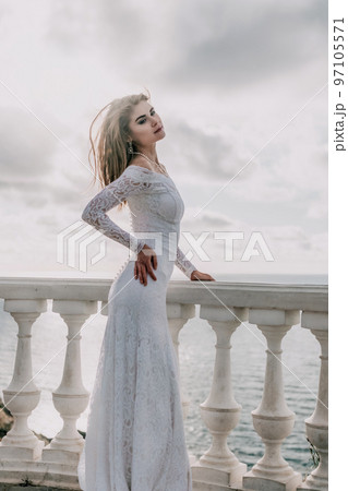 Romantic bride, blond girl in white wedding dress with open shoulders posing on open terrace with backdrop of the sea and rocks. Stylish young woman standing on terrace and looking on ocean at sunset 97105571