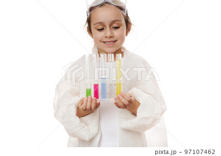 Selective focus on test tubes with colorful reagents and chemicals on tripod in the hands of a blurred child, little girl, young chemist scientist in lab coat, isolated on white background. Copy space 97107462