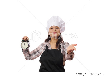 Female pastry chef in black apron, shows alarm clock with the opening time of a coffee shop or bakery, extends her hand, invite you to visit a new catering establishment, isolated on white background Female pastry chef in black apron, shows alarm clock with the opening time of a coffee shop or bakery, extends her hand, invite you to visit a new catering establishment, isolated on white background 97107490