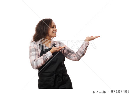 Pleasant cheerful Latin American woman waitress, barmaid, female bartender, barista wearing black chef's apron, smiles while pointing to copy ad space on her hand palm up, isolated on white background Pleasant cheerful Latin American woman waitress, barmaid, female bartender, barista wearing black chef's apron, smiles while pointing to copy ad space on her hand palm up, isolated on white background 97107495