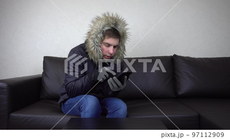 A young man is sitting at home in a winter jacket and trying to keep warm. A man sits without heating and calls on the phone and asks for help. A young man is sitting at home in a winter jacket and trying to keep warm. A man sits without heating and calls on the phone and asks for help. 97112909