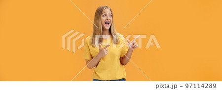 Amused enthusiastic and charismatic girl with blond hair and tanned pure skin without makeup pointing and looking at upper left corner with joyful happy smile over orange background 97114289