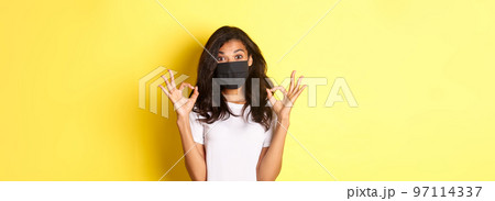 Concept of coronavirus, pandemic and lifestyle. Portrait of pleased african-american girl in black face mask, showing okay signs in approval, like something good, yellow background 97114337