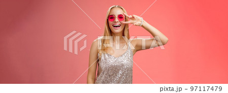 Energized friendly good-looking blond 25s woman in silver stylish dress sunglasses show peace victory gesture carefree smiling camera have fun enjoying cool party, standing red background 97117479