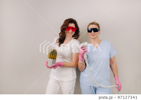Two cosmetologists in protective glasses are holding a laser depilation device and a cactus in their hands Two cosmetologists in protective glasses are holding a laser depilation device and a cactus in their hands 97127341