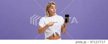 Waist-up shot of impressed and delighted gorgeous european female with blond hair and tattoos looking and pointing at awesome brand new smartphone standing over purple background Waist-up shot of impressed and delighted gorgeous european female with blond hair and tattoos looking and pointing at awesome brand new smartphone standing over purple background 97137717