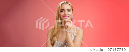 Waist-up shot elegant good-looking wealthy young blond woman in brilliant accessorize silver glittering stylish dress touch chin have clever interesting idea smiling curiously, red background 97137730