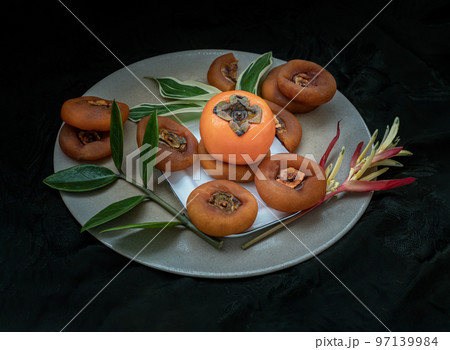 One fresh ripe persimmon fruit with Dried persimmons and colorful leaves on round ceramic tray. 97139984