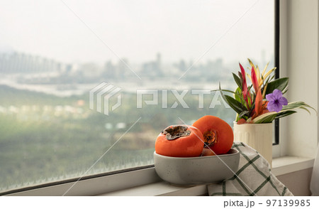 Few fresh ripe persimmons fruits in the ceramic bowl beside with Colorful flower vase. Few fresh ripe persimmons fruits in the ceramic bowl beside with Colorful flower vase. 97139985
