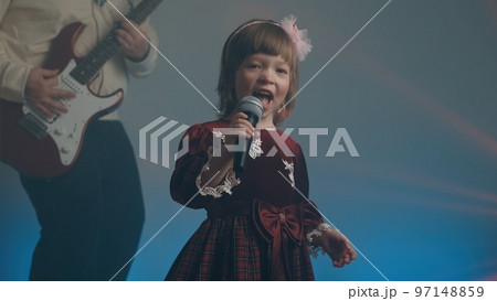 Little girl on stage in vintage dress, she sings into microphone and dances, her father plays an electric guitar. Color music is shining and smoke is billowing. Family time, creativity and hobbies 97148859
