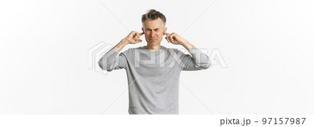 Portrait of annoyed and distressed middle-aged man, grimacing from loud disturbing noise, shut ears with fingers, standing over white background 97157987