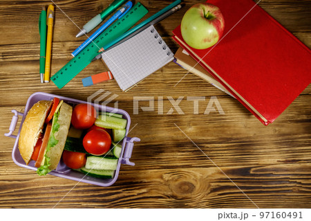 Back to school concept. School supplies, books, apple and lunch box with burgers and fresh vegetables on a wooden table. Top view 97160491