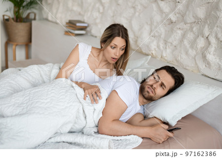 Shocked woman seeing her husband messaging to his lover on cellphone while lying in bed near him 97162386