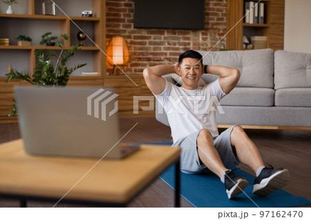 Smiling asian mature man working on abs, watching fitness tutorial on laptop and exercising on fitness mat at home 97162470
